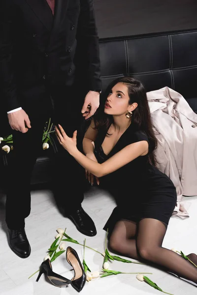 Attraktive Frau sitzt mit Tulpen auf dem Boden und umarmt stehenden Mann im Anzug in der Nähe der dunklen Couch — Stockfoto