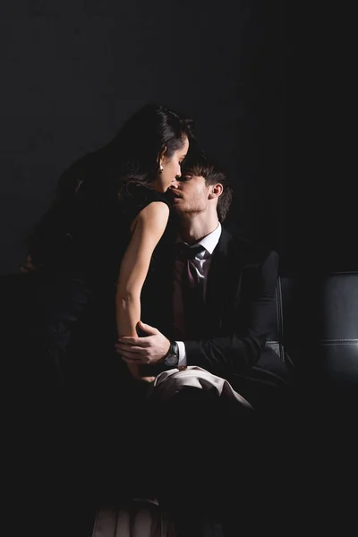 Man in suit and woman in black dress kissing on couch on dark background — Stock Photo