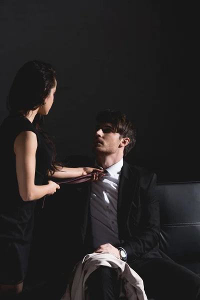 Brunetta donna in abito nero togliersi cravatta con bello seduto sul divano su sfondo scuro — Foto stock
