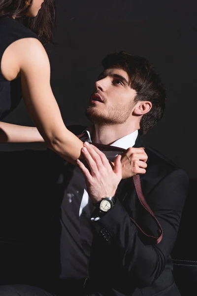 Frau im schwarzen Kleid zieht Krawatte aus, schöner Mann im Anzug sitzt auf Couch auf schwarzem Hintergrund — Stockfoto