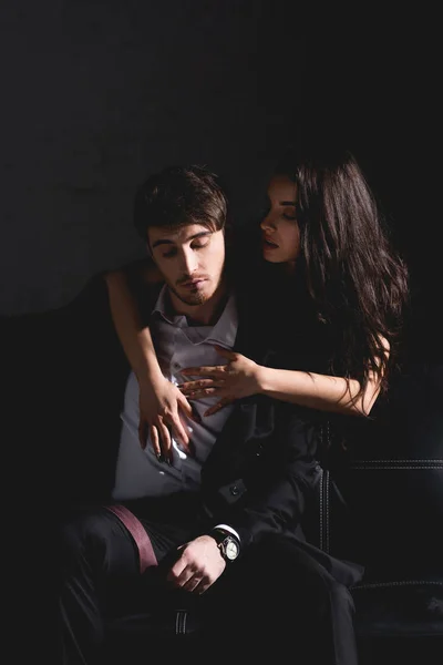 Brunette woman in black dress standing behind couch and hugging handsome man sitting on black background — Stock Photo