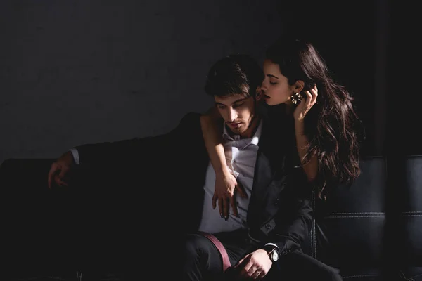 Brunette woman in black dress standing behind couch, hugging and kissing handsome man in suit sitting on black background — Stock Photo