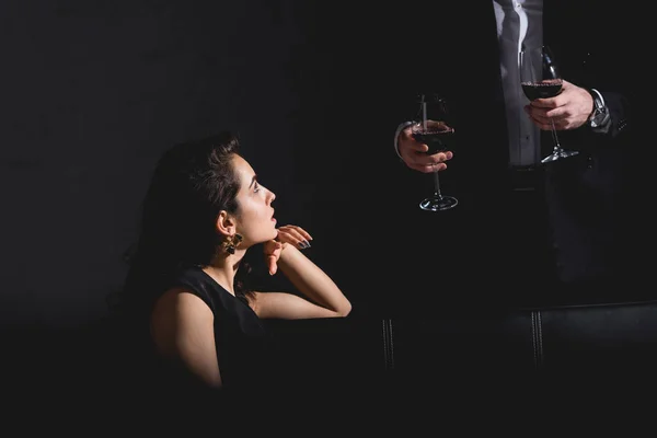 Atractiva mujer sentada en vestido negro astuto hombre guapo en traje de pie detrás del sofá y la celebración de vasos con vino sobre fondo negro - foto de stock