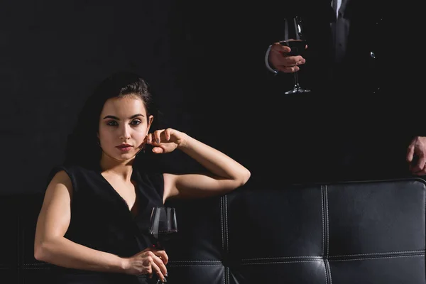 Attractive brunette woman sitting wile handsome man standing behind couch and holding glass with wine on black background — Stock Photo