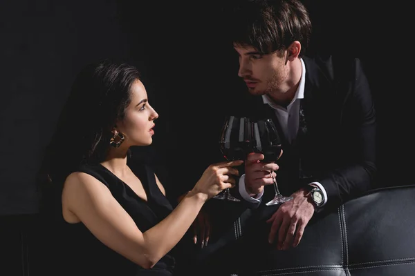 Attraktive Frau sitzt im Kleid und klirrt mit Brille, schöner Mann im schwarzen Anzug steht hinter Couch auf schwarzem Hintergrund — Stockfoto