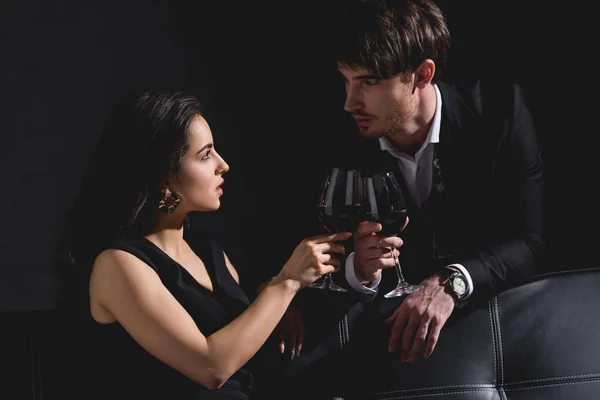 Atractiva mujer sentada en vestido y tintineo gafas con hombre guapo en traje negro de pie detrás del sofá sobre fondo negro - foto de stock