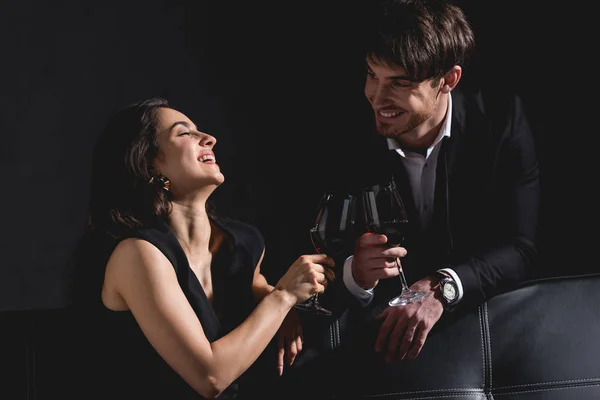 Donna attraente seduta in abito, occhiali di clinking con bell'uomo in piedi dietro il divano e sorridente su sfondo nero — Foto stock