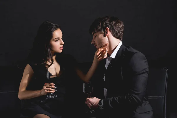 Atractiva mujer morena en vestido negro y hombre guapo en traje sentado en el sofá con vidrio sobre fondo negro - foto de stock