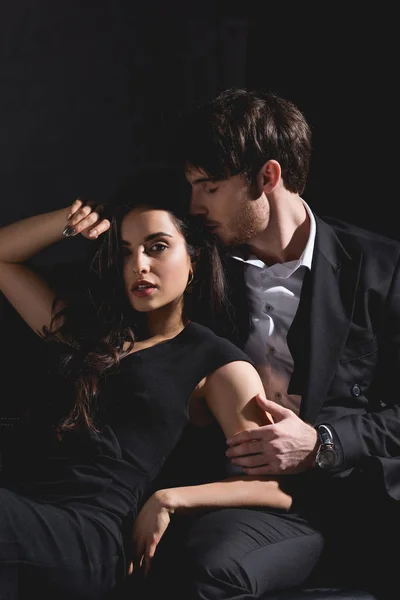 Atractiva mujer morena en vestido negro y hombre guapo en traje sentado en el sofá y abrazos aislados en negro - foto de stock