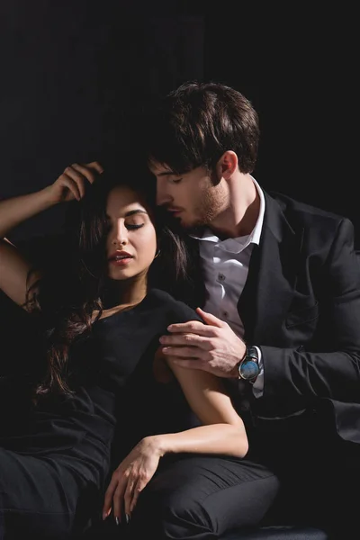 Hombre guapo en traje sentado en el sofá, abrazando y besando atractiva mujer morena en vestido negro aislado en negro - foto de stock