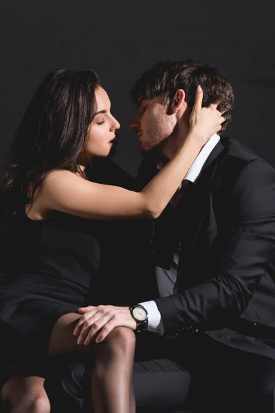 Man in suit and woman in black dress sitting and kissing on couch on black background — Stock Photo