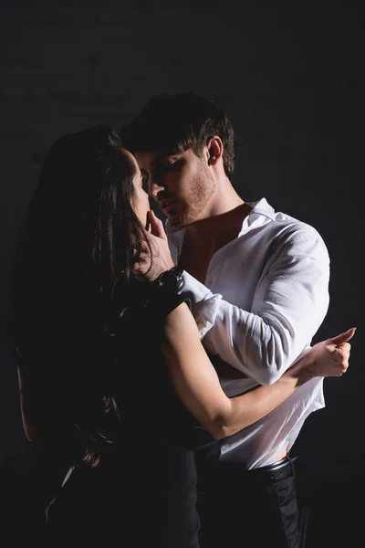 Brunetta donna in abito nero in piedi, baciare e spogliarsi uomo in camicia bianca su sfondo nero — Foto stock