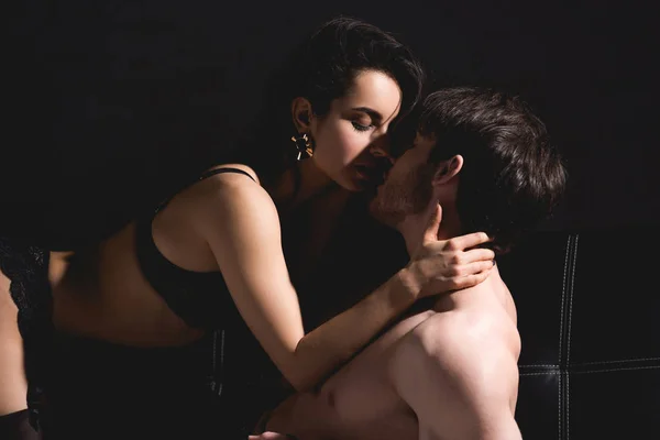 Sem camisa homem e mulher em lingerie preta beijando no sofá isolado no preto — Fotografia de Stock