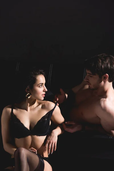 Handsome man lying on couch and undressing woman in lingerie sitting on floor on black background — Stock Photo