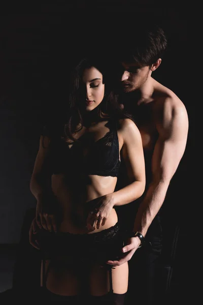 Attractive brunette woman in black lingerie and shirtless handsome man standing and hugging isolated on black — Stock Photo
