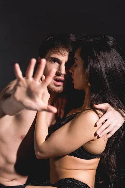 Woman in black lingerie hugging shirtless man showing stop sign with hand isolated on black — Stock Photo