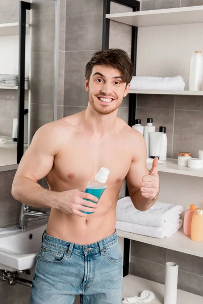 Hombre guapo sin camisa sonriendo, sosteniendo enjuague bucal y mostrando los pulgares hacia arriba con la mano en el baño - foto de stock