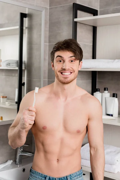 Bel homme torse nu souriant et tenant une brosse à dents dans la salle de bain — Photo de stock