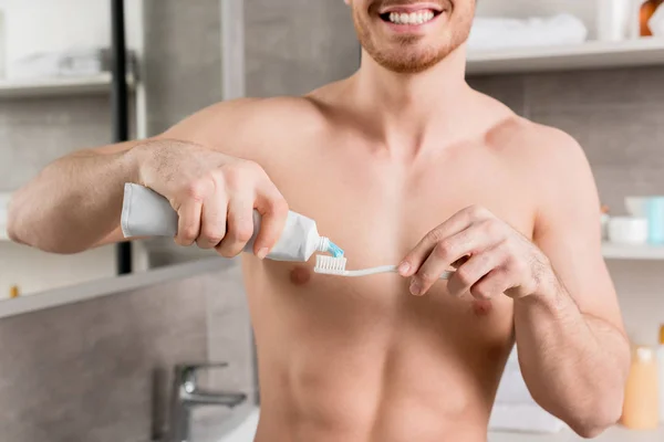 Vista recortada del hombre sin camisa sonriendo y poniendo pasta de dientes al cepillo de dientes en el baño - foto de stock