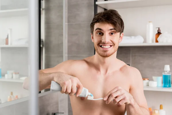 Красивий без сорочки чоловік посміхається і кладе зубну пасту зубну щітку у ванну — стокове фото