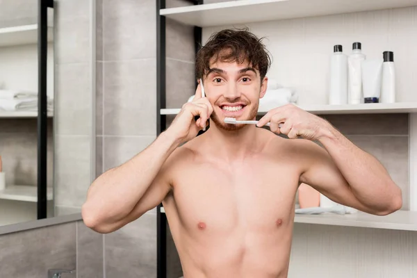 Homem sorrindo escovando os dentes e falando no smartphone no banheiro — Fotografia de Stock