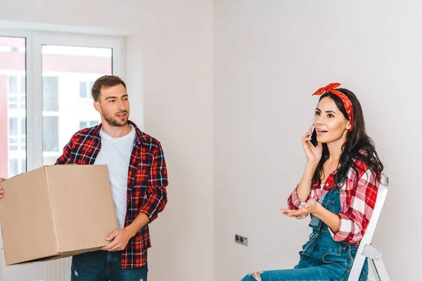 Homem bonito segurando caixa e olhando para a mulher falando no smartphone em casa — Fotografia de Stock