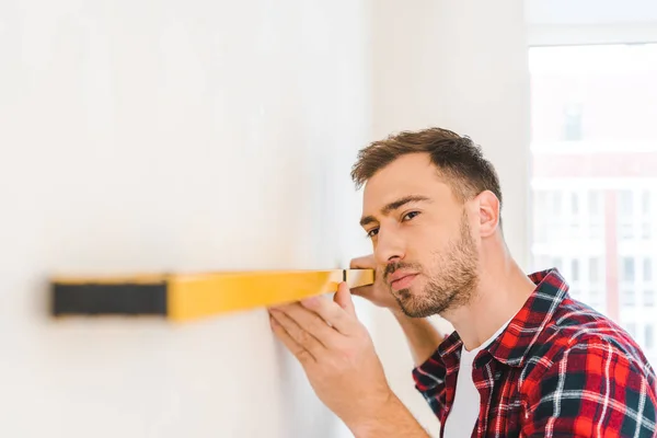 Handsome handyman holding measuring level while measuring wall at home — Stock Photo