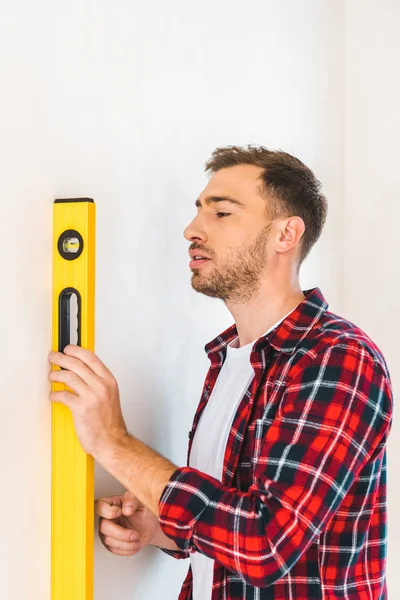 Homem bonito segurando nível de medição ao medir a parede em casa — Fotografia de Stock