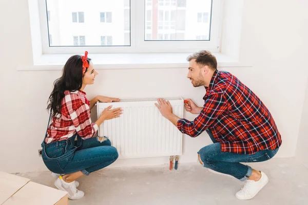 Fröhliches Paar berührt Heizkörper, während es am Fenster zu Hause sitzt — Stockfoto