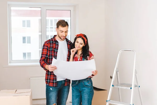 Couple joyeux debout près des boîtes et tenant le plan à la maison — Photo de stock