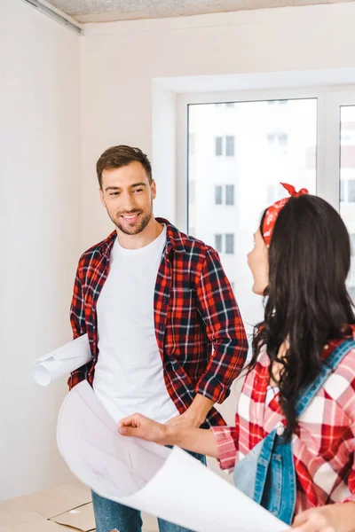 Selective focus of cheerful man looking at girlfriend holding blueprint at home — Stock Photo