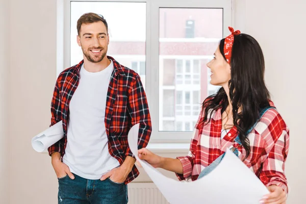 Cheerful man standing with hands in pockets and looking at girlfriend holding blueprint at home — Stock Photo