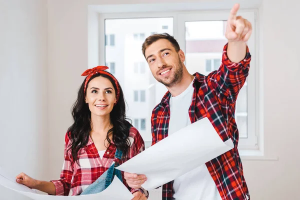 Hermosa mujer de pie con el hombre señalando con el dedo mientras sostiene el plano en casa - foto de stock