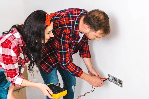 Cheerful woman holding digital multimeter near boyfriend looking at power socket — Stock Photo