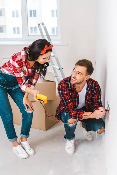 Attractive woman holding digital multimeter near cheerful boyfriend sitting near power socket and holding cables — Stock Photo