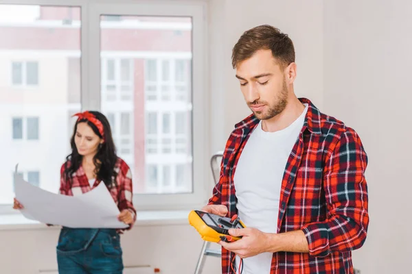 Selective focus of handsome man holding digital multimeter with woman holding blueprint on background — Stock Photo