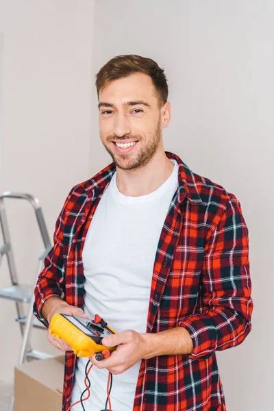 Homme joyeux tenant multimètre numérique dans les mains tout en se tenant à la maison — Photo de stock