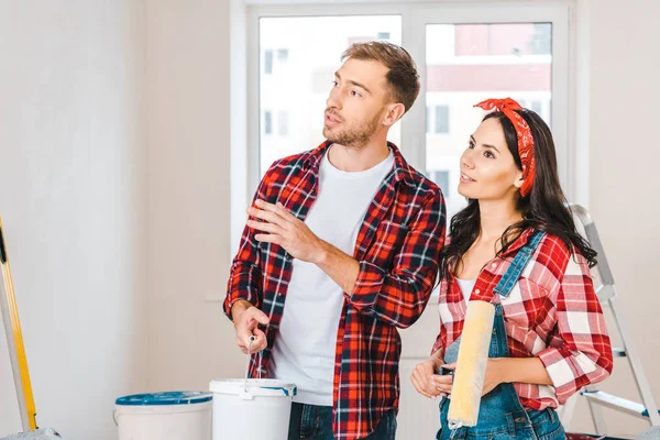 Feliz pareja sosteniendo cubo de pintura y rodillo mientras habla en casa - foto de stock