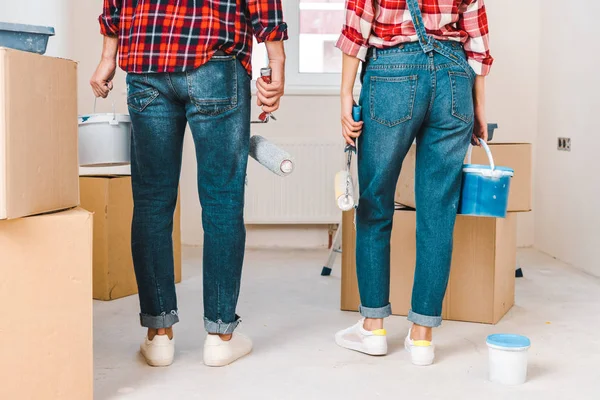 Vista trasera del hombre y la mujer sosteniendo cubos de pintura y rodillos mientras están de pie cerca de cajas - foto de stock