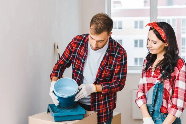 Homme tenant seau de peinture avec de la peinture bleue près de la femme portant des gants à la maison — Photo de stock