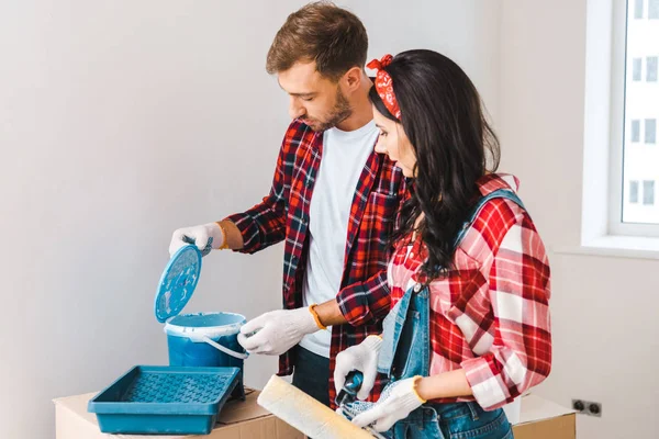 Handsome man opening paint bucket with blue paint near woman wearing gloves at home — Stock Photo