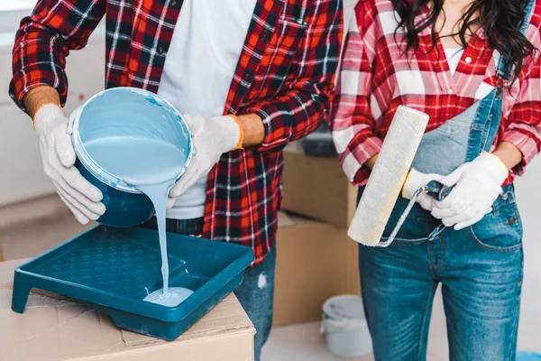 Vista cortada do homem derramando balde de pintura com tinta azul perto de mulher segurando rolo — Fotografia de Stock