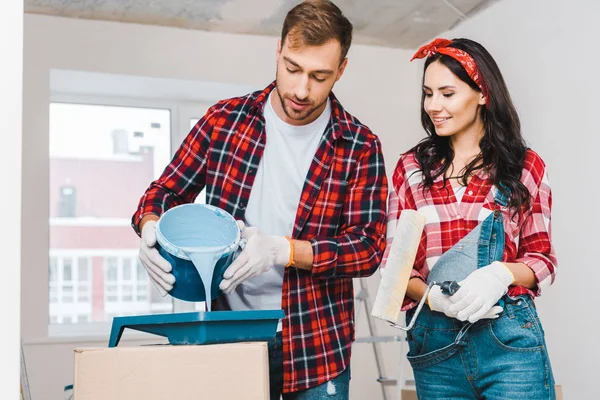 Homem bonito derramando balde de pintura com tinta azul perto de mulher alegre segurando rolo — Fotografia de Stock