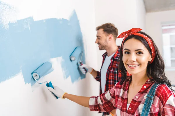 Enfoque selectivo de mujer atractiva sosteniendo rodillo cerca de hombre pintura pared - foto de stock