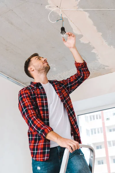 Schöner Mann wechselt Glühbirne, während er auf Leiter steht — Stockfoto