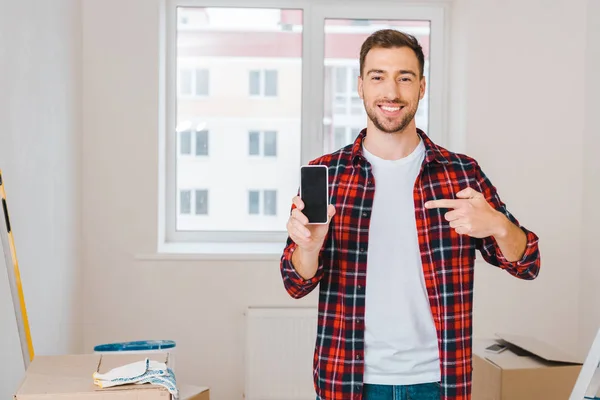 Веселий чоловік вказує пальцем на смартфон з порожнім екраном, стоячи вдома — стокове фото