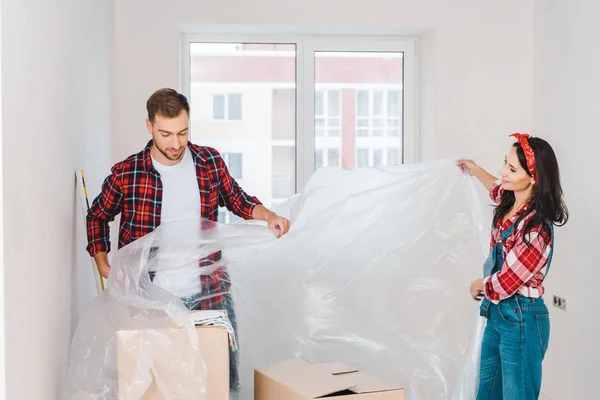 Alegre pareja cubriendo habitación con polietileno en casa - foto de stock