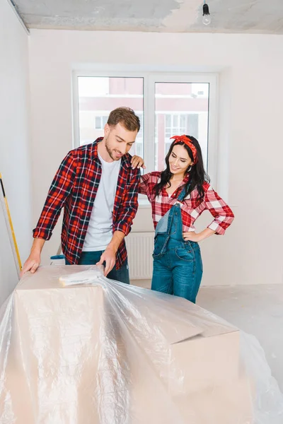 Cheerful couple looking at boxes covered with polyethylene at home — Stock Photo