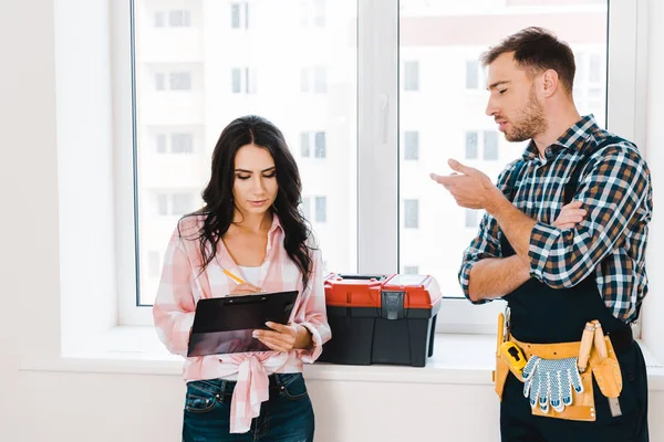 Attractive woman holding clipboard near handyman with tool belt on waist — Stock Photo