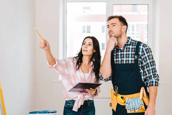 Attractive woman holding clipboard and pointing with finger near handyman — Stock Photo